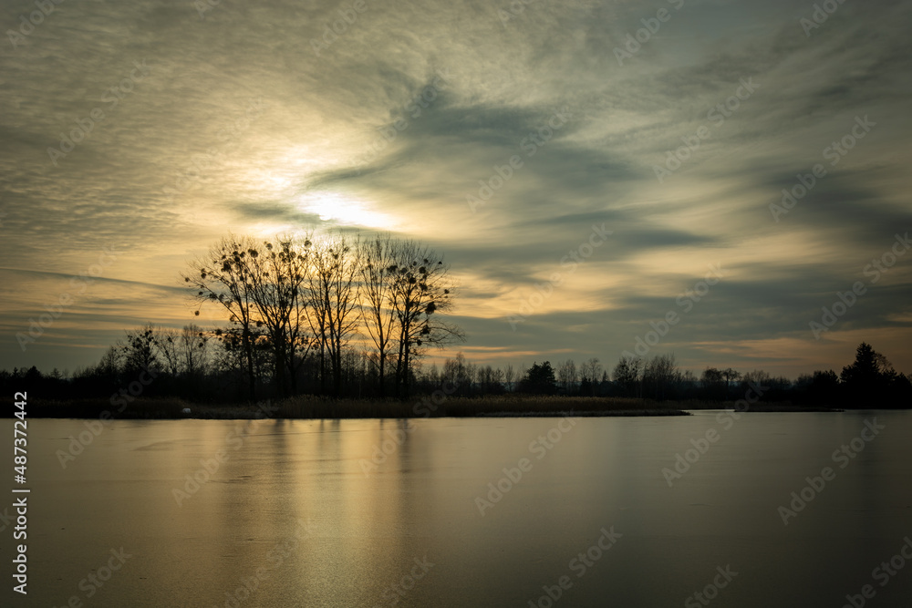 The sun behind the clouds over the frozen lake and trees on the shore