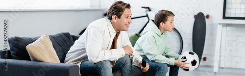 Man with remote controller smiling while watching football match with son at home, banner