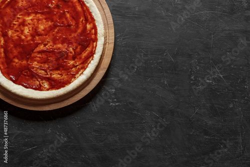 Round pizza dough spread with tomato sauce, preparation for cooking italian dish, top view dark background, with space to copy text.