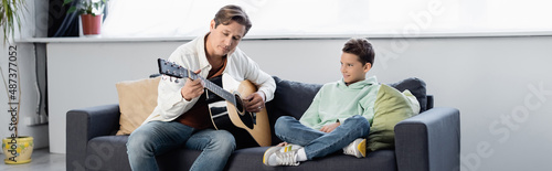 Father playing acoustic guitar near preteen son on couch, banner