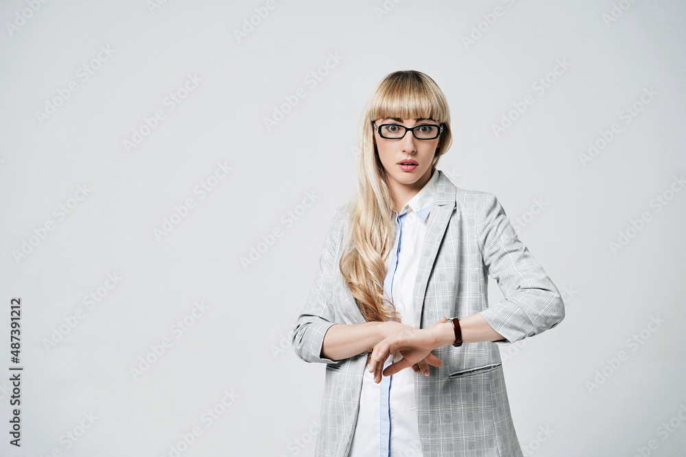 Young beautiful blonde woman in studio on light gray background. Model in pantsuit and glasses checking time with a wristwatch.