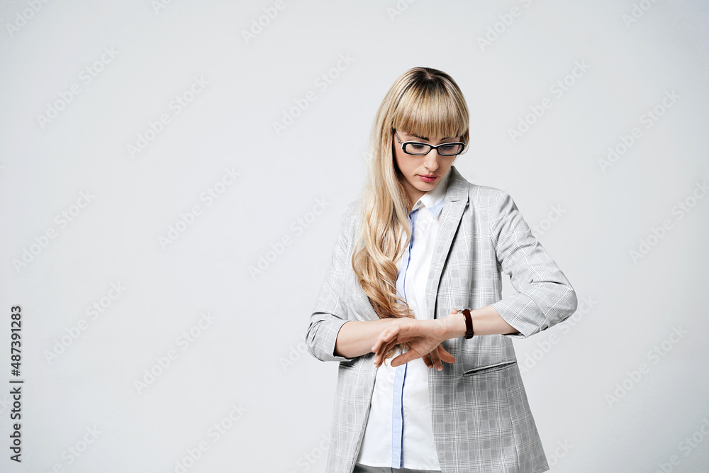 Young beautiful blonde woman in studio on light gray background. Model in pantsuit and glasses checking time with a wristwatch.
