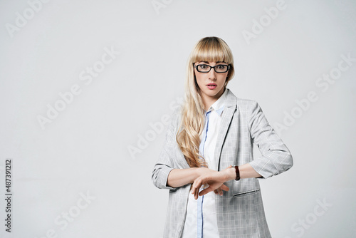Obraz na płótnie Young beautiful blonde woman in studio on light gray background. Model in pantsuit and glasses checking time with a wristwatch. w salonie