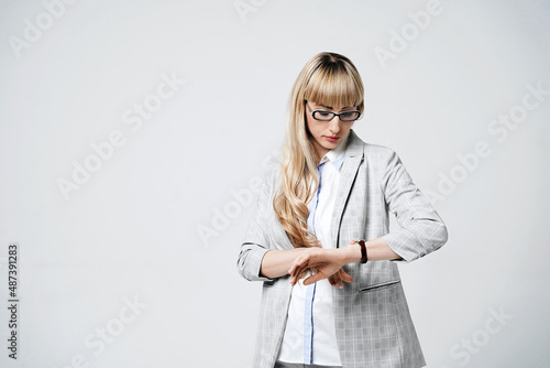 Young beautiful blonde woman in studio on light gray background. Model in pantsuit and glasses checking time with a wristwatch.