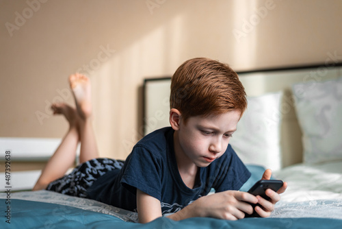 Communication and technology concept - ginger boy thinking with smartphone, texting or playing game at home