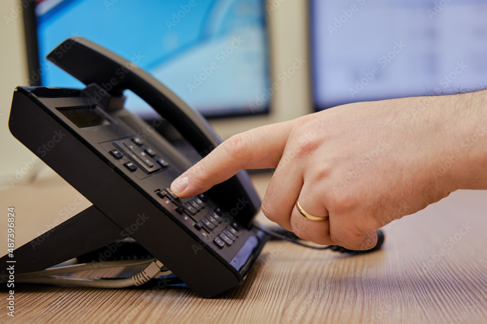 A man hand dials a number on the phone at the monitors of office computers. Finger presses on the buttons of a office landline phone A man hand dials a number on the phone at the monitors of office computers. Finger presses on the buttons of a office landline phone