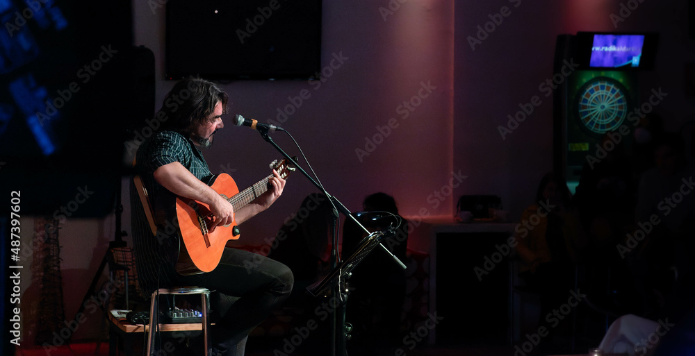 Fototapeta premium Singer-songwriter plays guitar and sings at acoustic concert in a club with an intimate atmosphere. Silhouettes of the public in the background