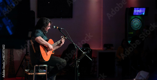 Singer-songwriter plays guitar and sings at acoustic concert in a club with an intimate atmosphere. Silhouettes of the public in the background