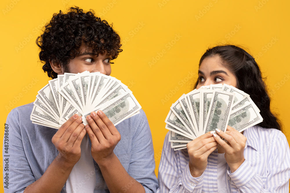 Big win concept. Young indian couple holding and covering faces with a lot of dollar cash fan, yellow studio background Big win concept. Young indian couple holding and covering faces with a lot of dollar cash fan, yellow studio background