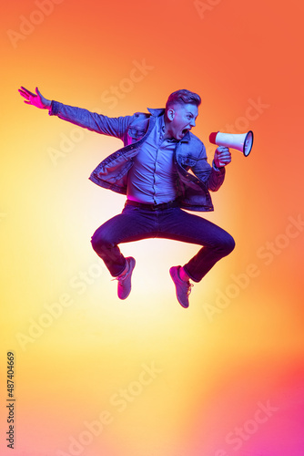 Full-length portrait of young excited man jumping with megaphone isolated on orange background in neon light, filter. Concept of emotions, beauty, fashion