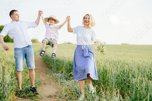 Happy family running together holding hands in summer. Teamwork. Family weekend. Mom, dad and son are playing on a green wheat field, a happy kid in straw hat holding parents' hands and jumping.