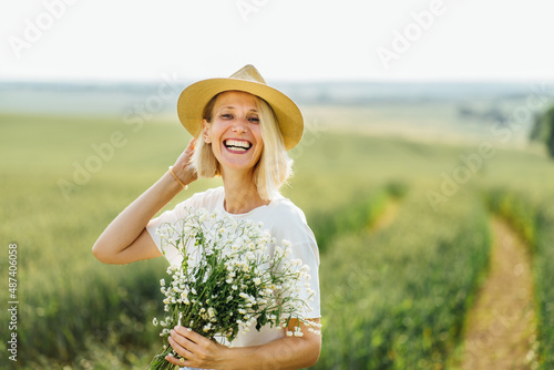 Gorgeous middle age blond woman with a bouquet of wild flowers in a wheat field on a sunset. A fashionable slim female in sraw hat rejoices, laughs, enjoys life and summer, nature, happiness.