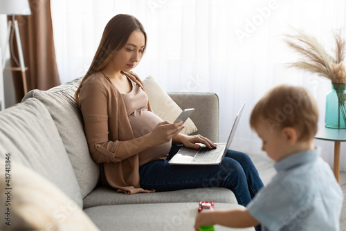Young pregnant freelancer woman using smartphone and laptop, mom taking care of her toddler son