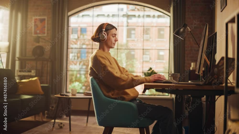 Young Handsome Man Working from Home on Desktop Computer in Sunny Stylish Loft Apartment. Creative Male Checking Social Media, Browsing Internet. Urban City View from Big Window. Handheld Shot. Young Handsome Man Working from Home on Desktop Computer in Sunny Stylish Loft Apartment. Creative Male Checking Social Media, Browsing Internet. Urban City View from Big Window. Handheld Shot.