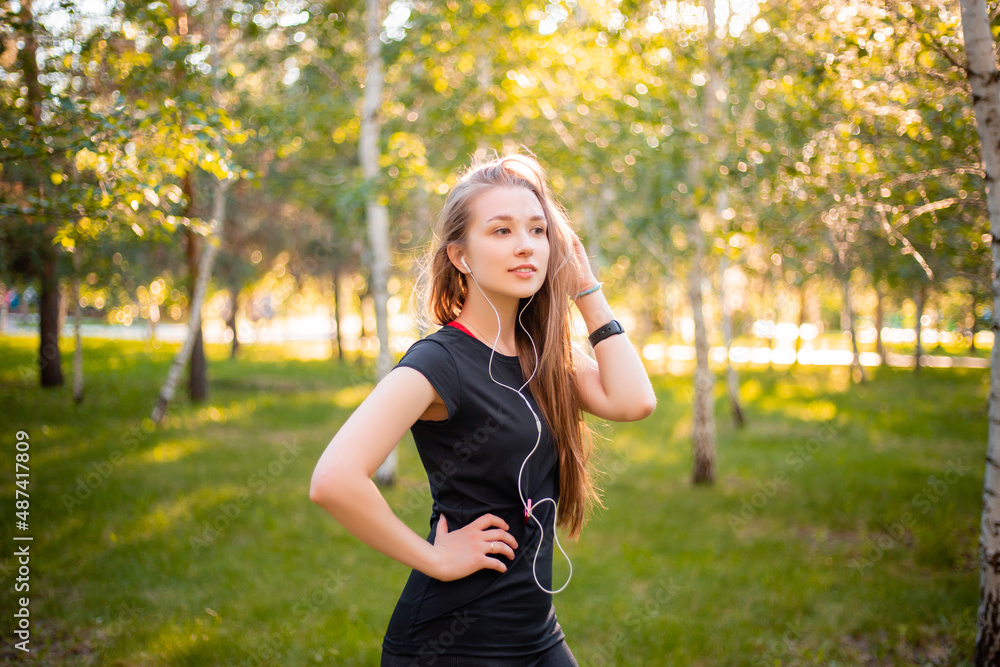 Portrait of a beautiful smiling girl in a sports uniform with flowing hair listening to music in headphones on the background of the park. Portrait of a beautiful smiling girl in a sports uniform with flowing hair listening to music in headphones on the background of the park.