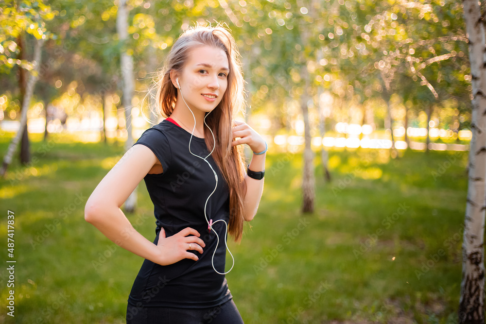Portrait of a beautiful smiling girl in a sports uniform with flowing hair listening to music in headphones on the background of the park. Portrait of a beautiful smiling girl in a sports uniform with flowing hair listening to music in headphones on the background of the park.