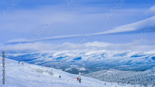 View of ski slope in Colorado ski resort; several skiers ski down the slope; beautiful view of mountain range and blue sky in background