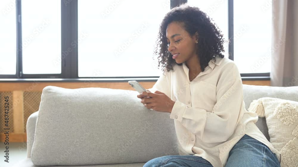 Cheerful young african woman using smartphone while sitting on couch. Black smiling woman using app on cellphone at home. Beautiful girl relaxing while chatting on mobile phone and looking at camera. Cheerful young african woman using smartphone while sitting on couch. Black smiling woman using app on cellphone at home. Beautiful girl relaxing while chatting on mobile phone and looking at camera.