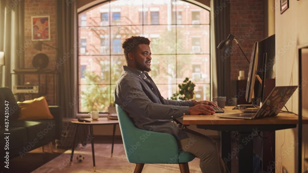 Young Handsome African American Man Working from Home on Desktop Computer in Sunny Stylish Loft Apartment. Creative Male Checking Social Media, Browsing Internet. Urban City View from Big Window. Young Handsome African American Man Working from Home on Desktop Computer in Sunny Stylish Loft Apartment. Creative Male Checking Social Media, Browsing Internet. Urban City View from Big Window.