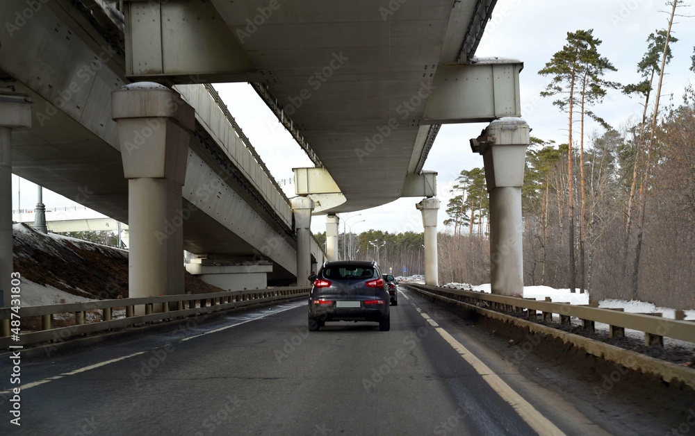 Cars are moving in a traffic jam under a trestle bridge in a forest belt with tall pines. Cars are moving in a traffic jam under a trestle bridge in a forest belt with tall pines.
