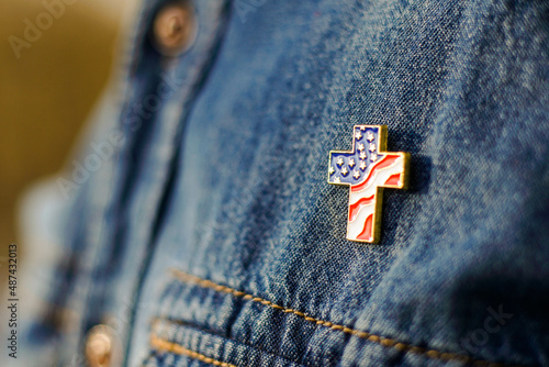 Close up of Christian cross pin with American flag colors is pinned on blue jeans jacket. Patriotism and religious rights concept