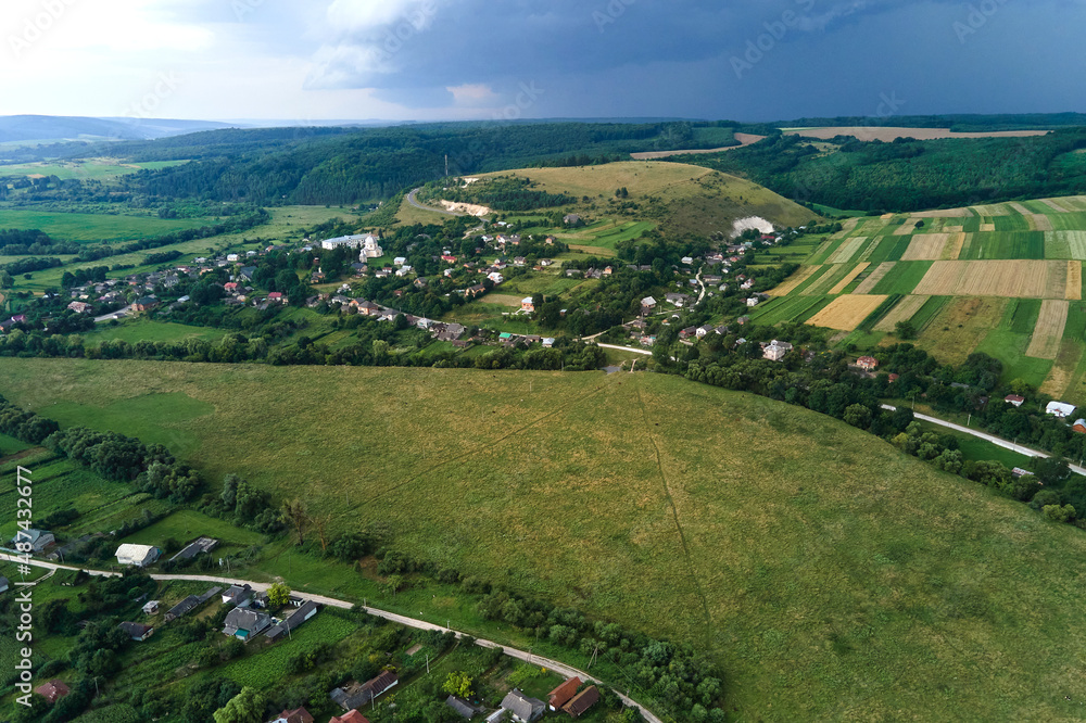 Aerial landscape view of village houses and distant green cultivated agricultural fields with growing crops on bright summer day