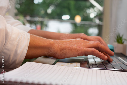 hands of a woman dressed in a white shirt working or studying on her computer from home and in the background you can see the green plants of her garden photo