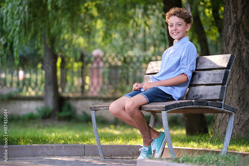 Young happy child boy relaxing sitting on bench in summer park. Positive kid enjoying summertime outdoors. Child wellbeing concept