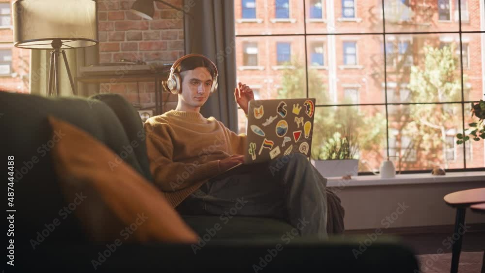 Young Handsome Man Sitting on Sofa and Using Laptop Computer in Sunny Stylish Loft Apartment. Creative Designer Wearing Cozy Yellow Sweater and Headphones. Urban City View from Big Window. Young Handsome Man Sitting on Sofa and Using Laptop Computer in Sunny Stylish Loft Apartment. Creative Designer Wearing Cozy Yellow Sweater and Headphones. Urban City View from Big Window.