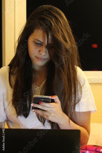 Photo of a young, smiling woman has a video call at night while lying on a bed in her bedroom photo