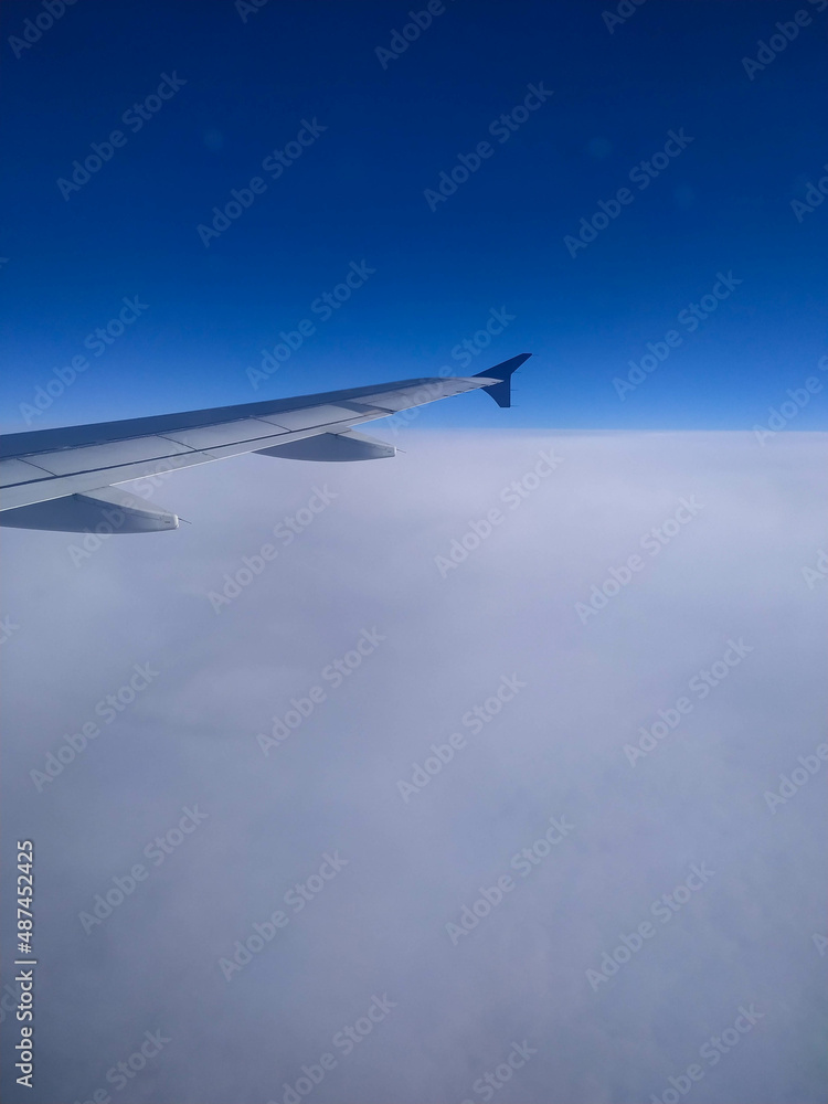 Fototapeta premium The view from the plane window on a cloudy sky in winter. Cabin window, snow, blue sky, plane wing, frozen plane window, flying. View on earth from a plane.