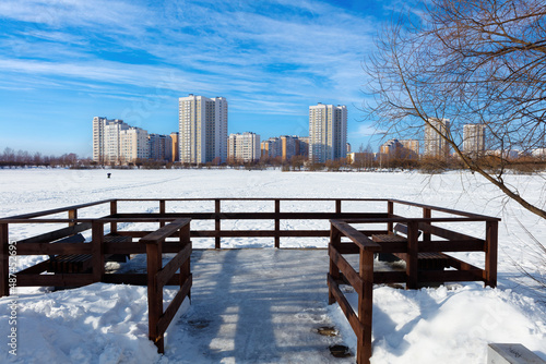 Winter ice fishing, lake, frosty day. A fisherman is engaged in ice fishing in the pond of the city park. Against the background of a multi-storey, modern, residential development.