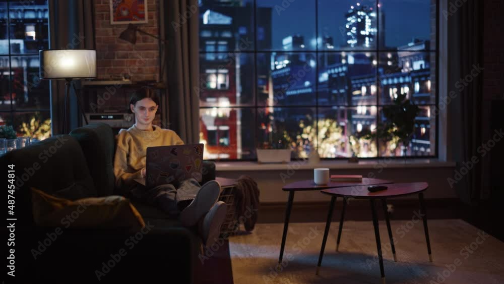 Young Handsome Man Working from Home on Laptop Computer in Stylish Loft Apartment in the Evening. Creative Person Checking Social Media, Browsing Internet. Urban City View from Big Window.