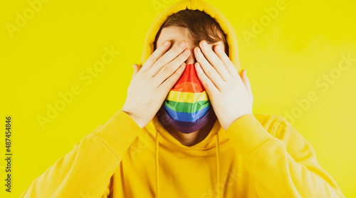 Scared man in mask lgbt covering face with hands. Young male in yellow sweatshirt and mask covering eyes with hands on yellow background