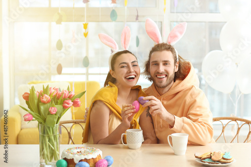 Canvas Print Happy couple in bunny ears cracking Easter eggs at table