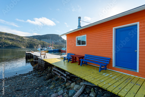 Fotografija The exterior wall of an orange color wooden boathouse with a vibrant blue door, closed glass window, blue wooden benches, and long wharf with boats