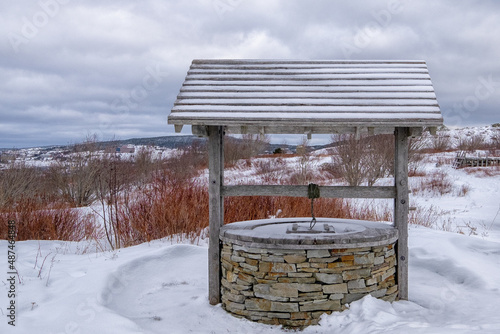 A wishing well with a rock base and a wooden roof. The crank is wood and the rotating piece is wooden with rope. The well is on snow covered hill with a grey cloudy sky. There are shrubs and trees. photo