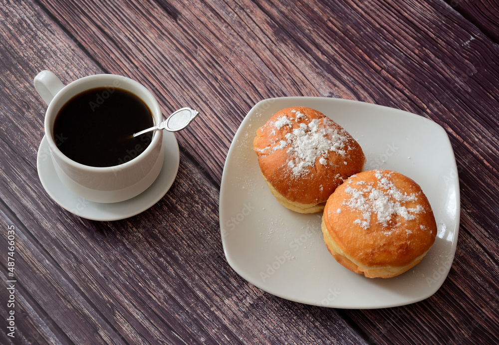 A cup of fresh black coffee and a plate with two donuts in powdered sugar on a wooden table. A cup of fresh black coffee and a plate with two donuts in powdered sugar on a wooden table.