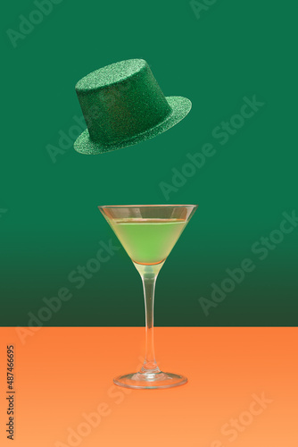 Irish festival minimal concept. Martini glass with green cocktail on terracotta table. Green glitter hats levitate above on a dark green background.
