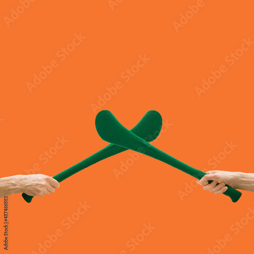Man hands hold criss-crossed two hurly sticks – hurleys in green color with shamrock decoration.  Creative copy space above against an orange background