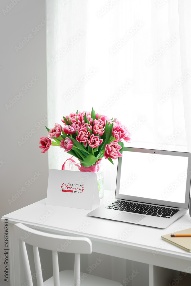 Workplace with modern laptop, tulips and greeting card with text HAPPY WOMAN'S DAY in light room Workplace with modern laptop, tulips and greeting card with text HAPPY WOMAN'S DAY in light room