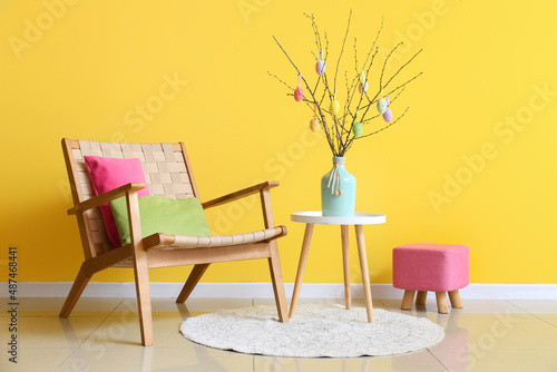 Tree branches with Easter eggs in vase on table, armchair and pouf near yellow wall