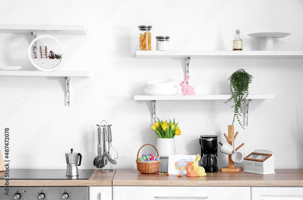 Counter with beautiful flowers, basket of Easter eggs, decor and different utensils near light wall Counter with beautiful flowers, basket of Easter eggs, decor and different utensils near light wall