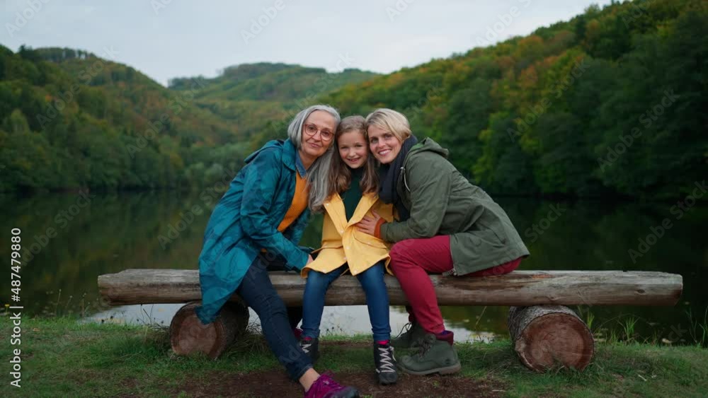 Small girl with mother and grandmother sitting on bench and looking at camera outoors by lake. Small girl with mother and grandmother sitting on bench and looking at camera outoors by lake.