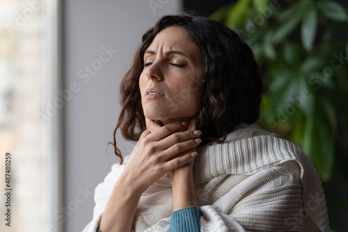Upset sick woman touching painful swollen neck glands, suffering from throat infection, unhealthy female standing near window at home feeling pain with swallowing, selective focus photo