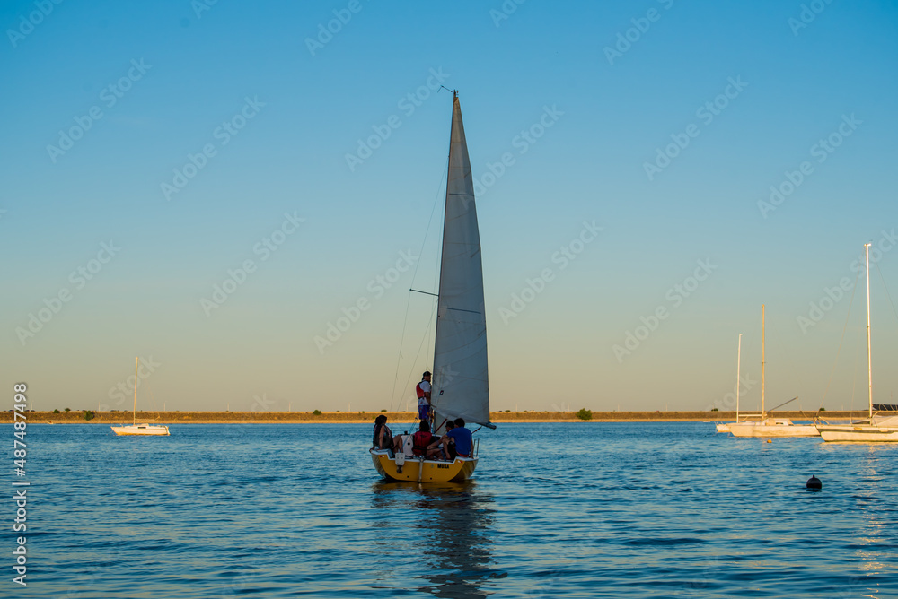 small sailboat over the water with people inside, boats and sky in the background