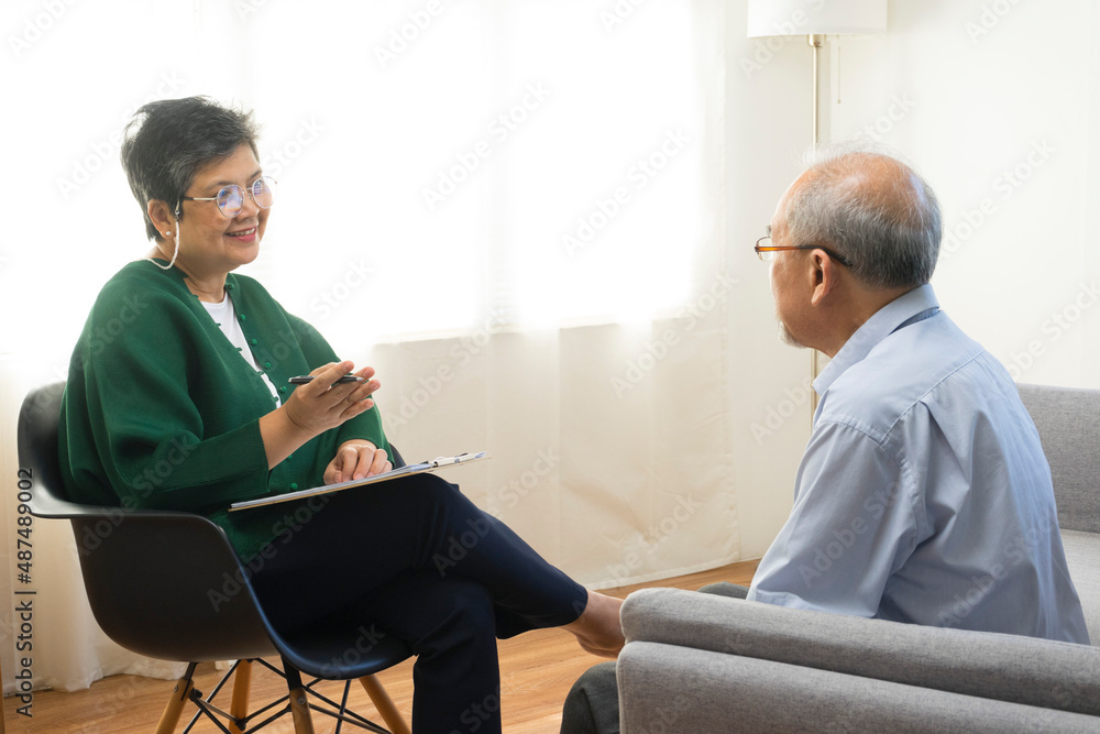 Depressed Asian senior man sharing problems during therapy session with female psychiatrist writing on clipboard. Depressed Asian senior man sharing problems during therapy session with female psychiatrist writing on clipboard.