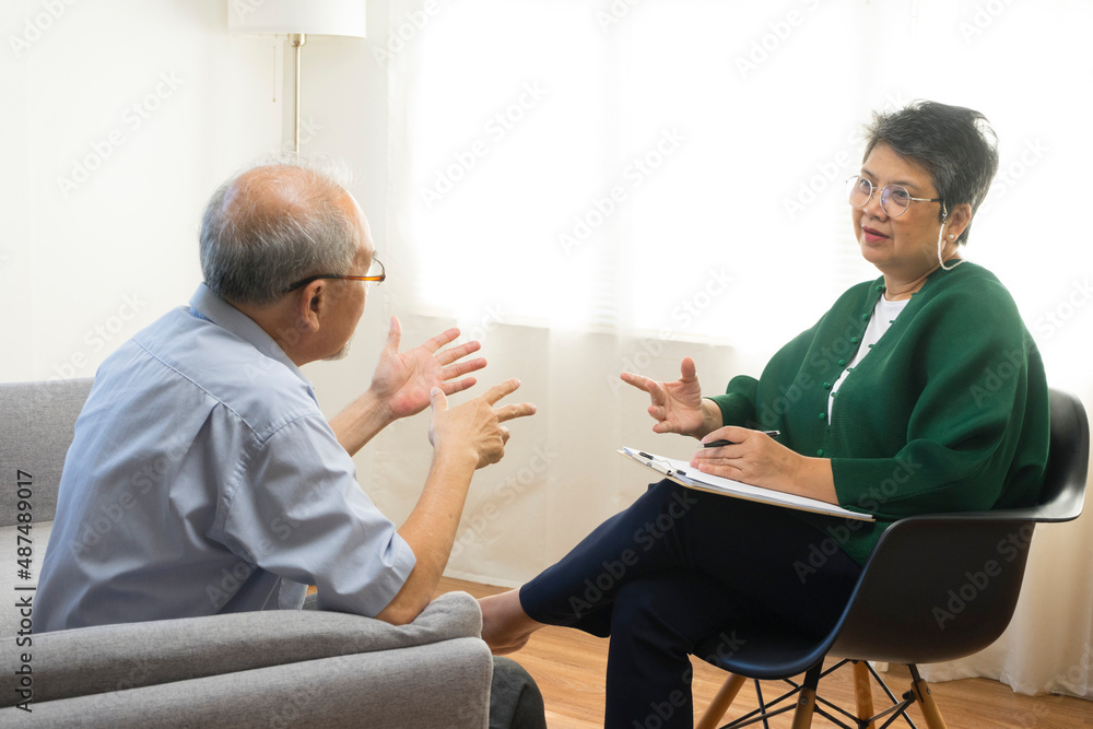 Depressed Asian senior man sharing problems during therapy session with female psychiatrist writing on clipboard. Depressed Asian senior man sharing problems during therapy session with female psychiatrist writing on clipboard.
