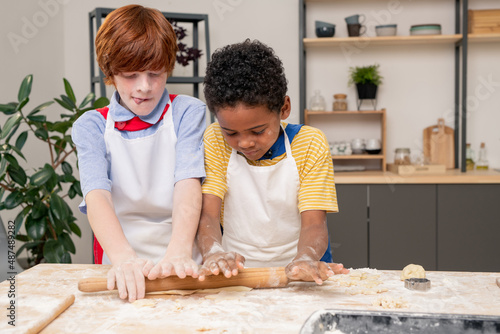 Cute blond boy rolling homemade dough on wooden table with his mom standing near by while cooking together