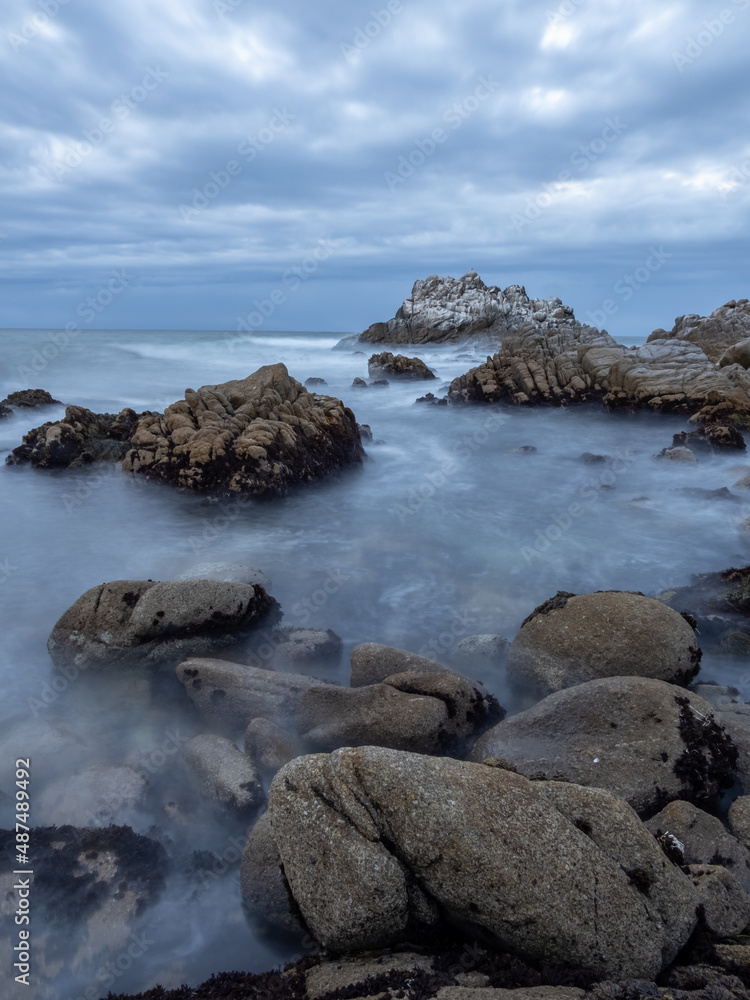 A rocky section of coastline near Monterey Bay in California, on an overcast day at sunset, long exposure.